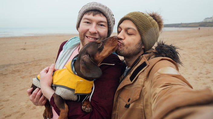 A couple in beanies on the beach holding their Dachshund dog who is wearing a yellow rain slicker. The dog is licking on one person's face.