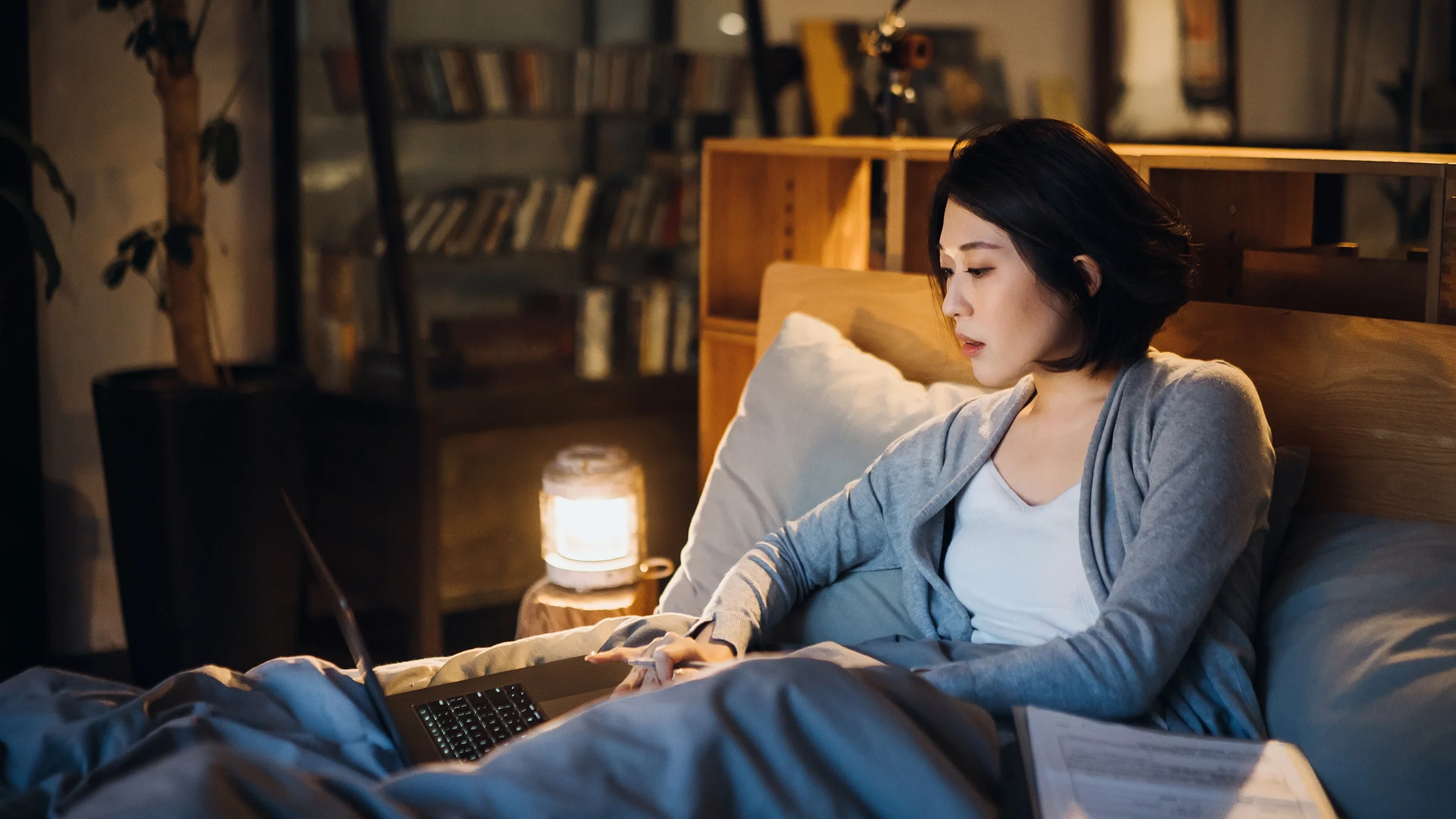 Young woman working from bed. She is working on her laptop and has a pile of paperwork next to her.