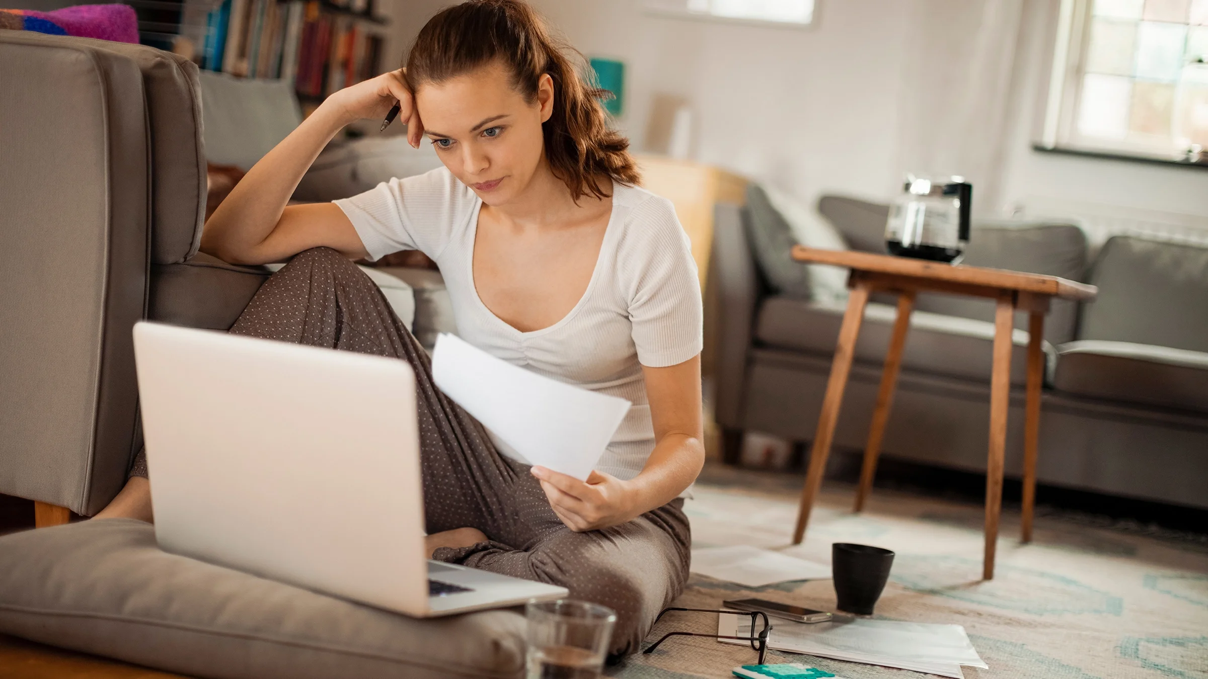 A woman sits on the floor of her living room. She leans against her couch while working on a laptop.