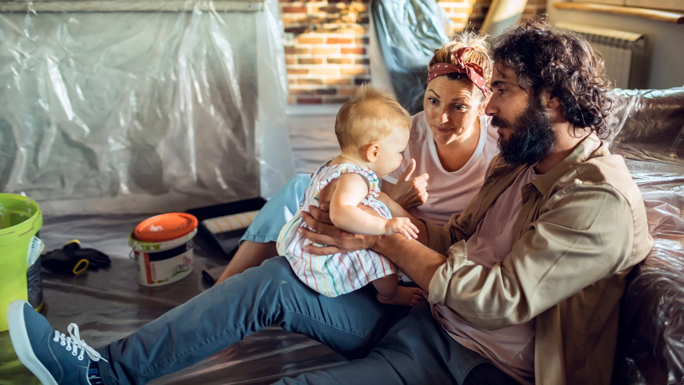 A family with a new baby sits in their living room, which is under renovation. There is plastic covering the furniture and paint supplies all around.