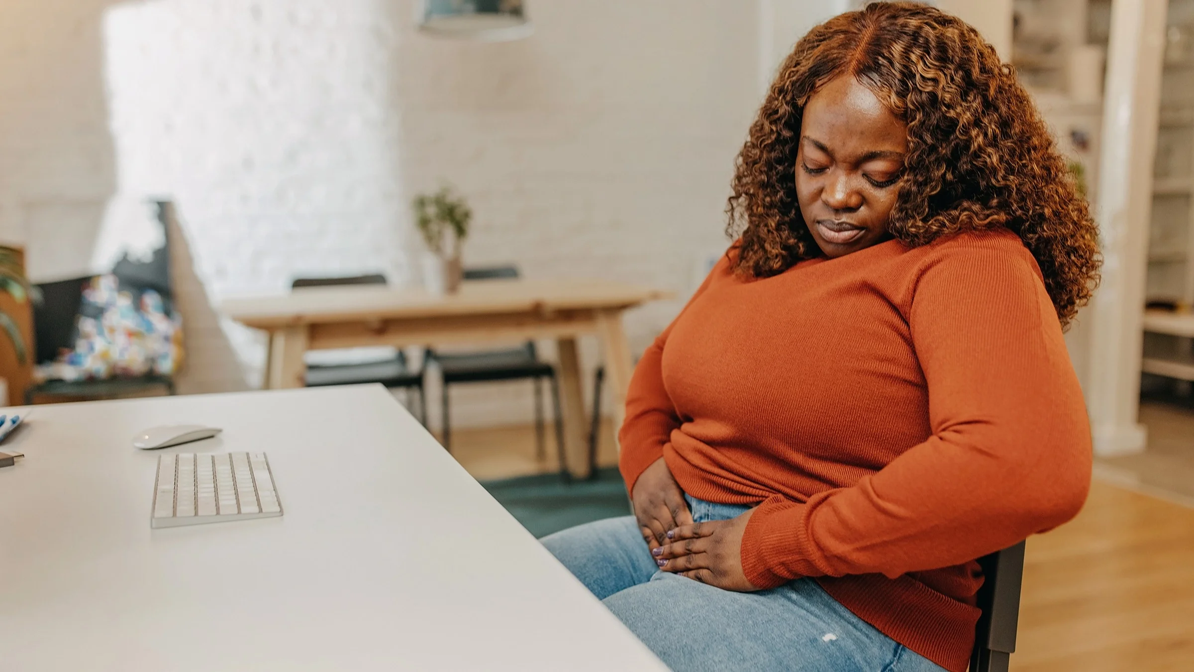 A woman with abdominal pain is sitting at her desk at home. She is wearing an orange long-sleeve sweater.