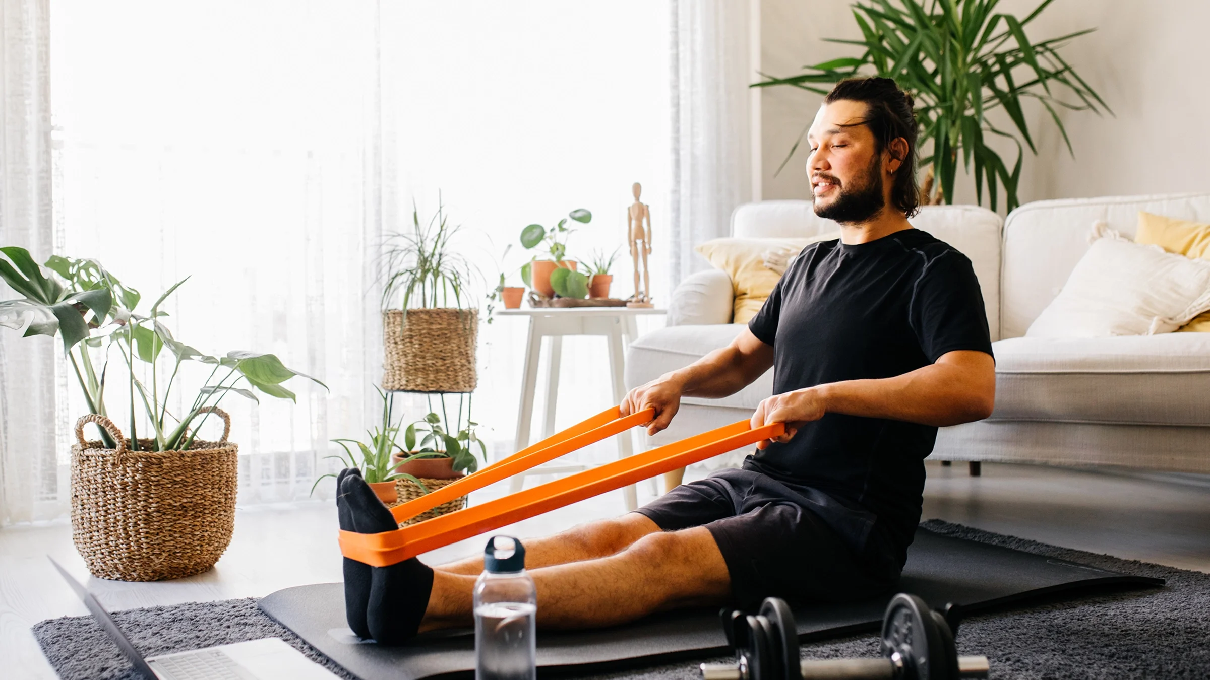 A man sitting on a yoga mat at home is exercising with resistance bands. 