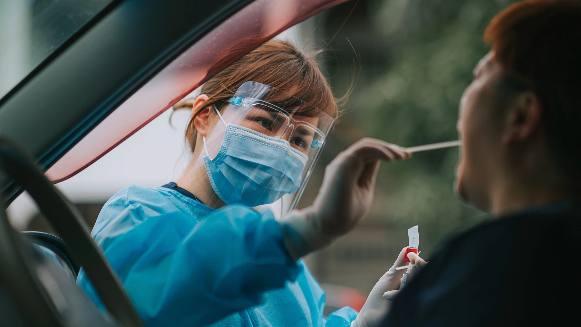Woman getting drive-thru COVID-19 test in car administered by a healthcare worker in blue scrubs, face mask, and face shield
