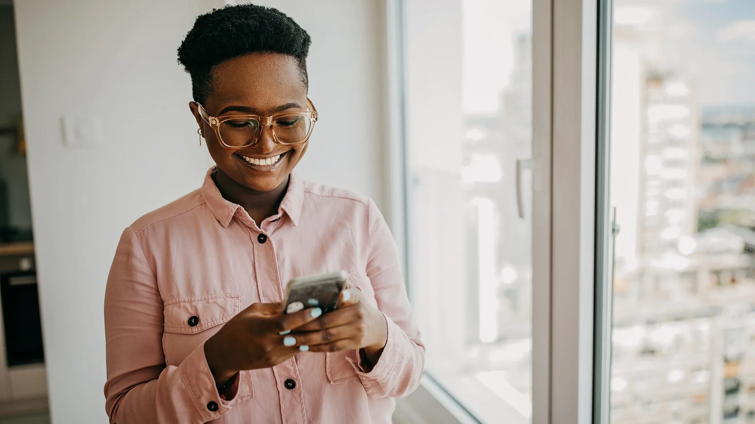 Young woman with short hair using her mobile phone and smiling. She is standing next to a window inside of a high rise building.
supersizer/E+ via Getty Images