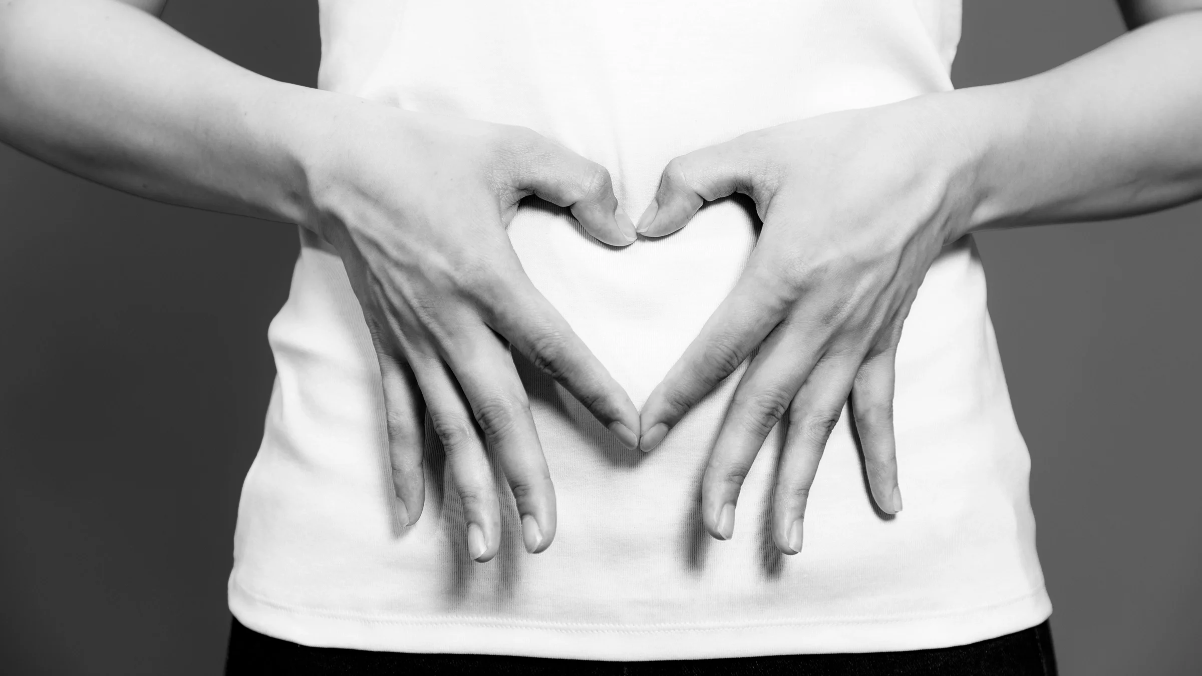 A cropped image in black and white shows a person making a heart with their hands on their stomach.