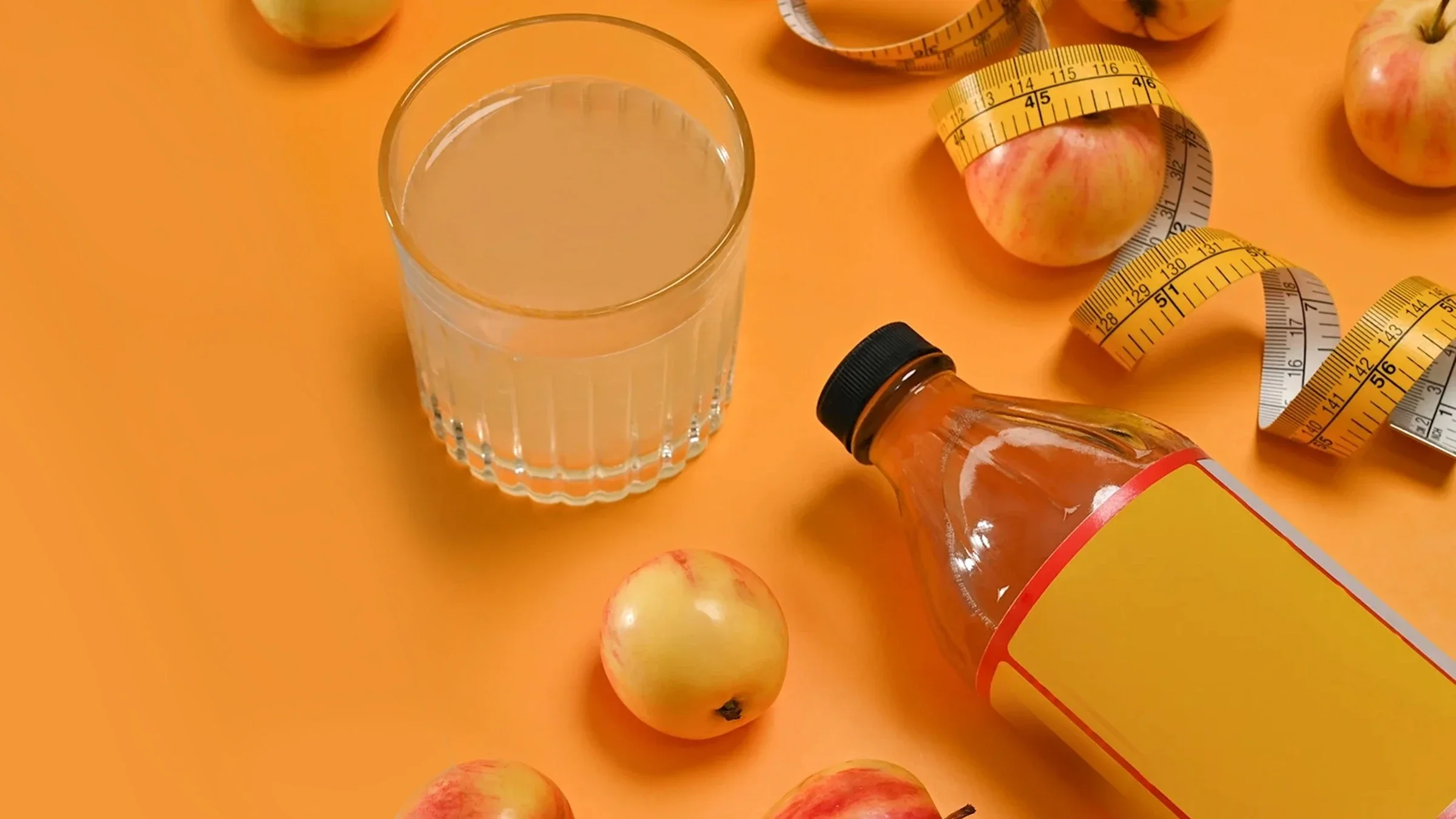 A glass of apple cider vinegar, bottle of apple cider vinegar, measuring tape, and apples on a table.