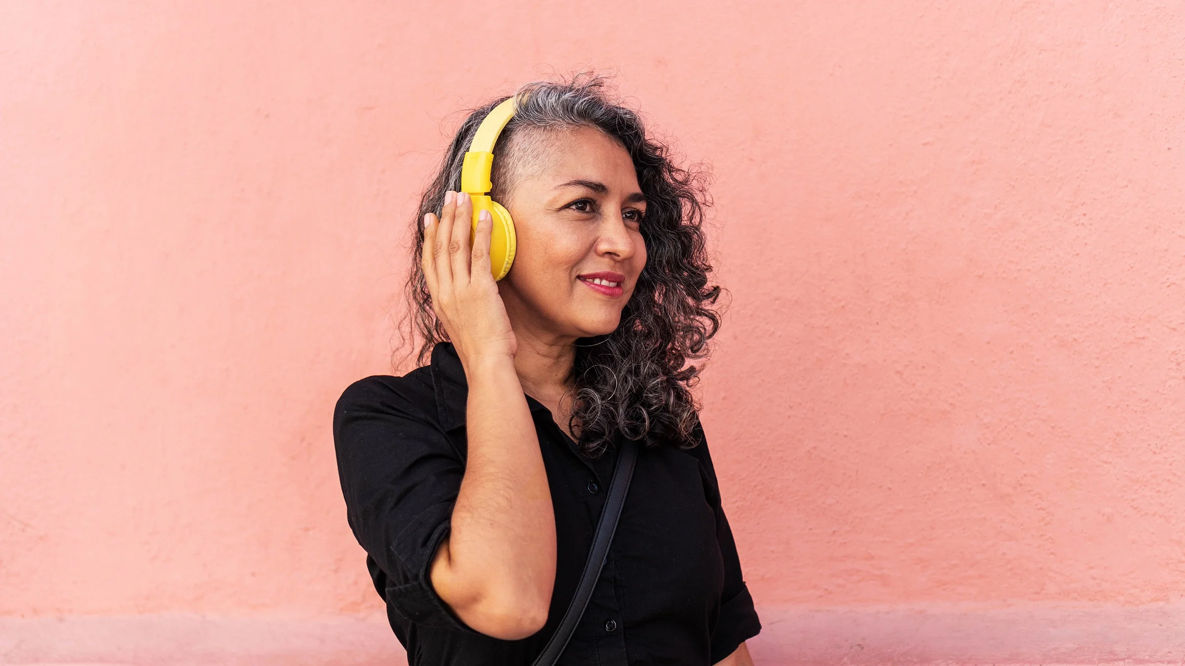A woman wearing yellow headphones stands before a pink wall.