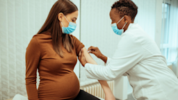 Pregnant woman at the doctor's office. The doctor is prepping her arm for a vaccine. Both are wearing blue medical face masks.
bogdankosanovic/E+ via Getty Images