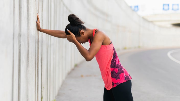 Young woman taking a break on her run. She has her head in one hand and is resting the other on a wall. She is wearing a bright pink workout top.