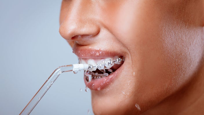 A close-up image shows a smiling woman with braces as she cleans her teeth and gums with a water flosser.
