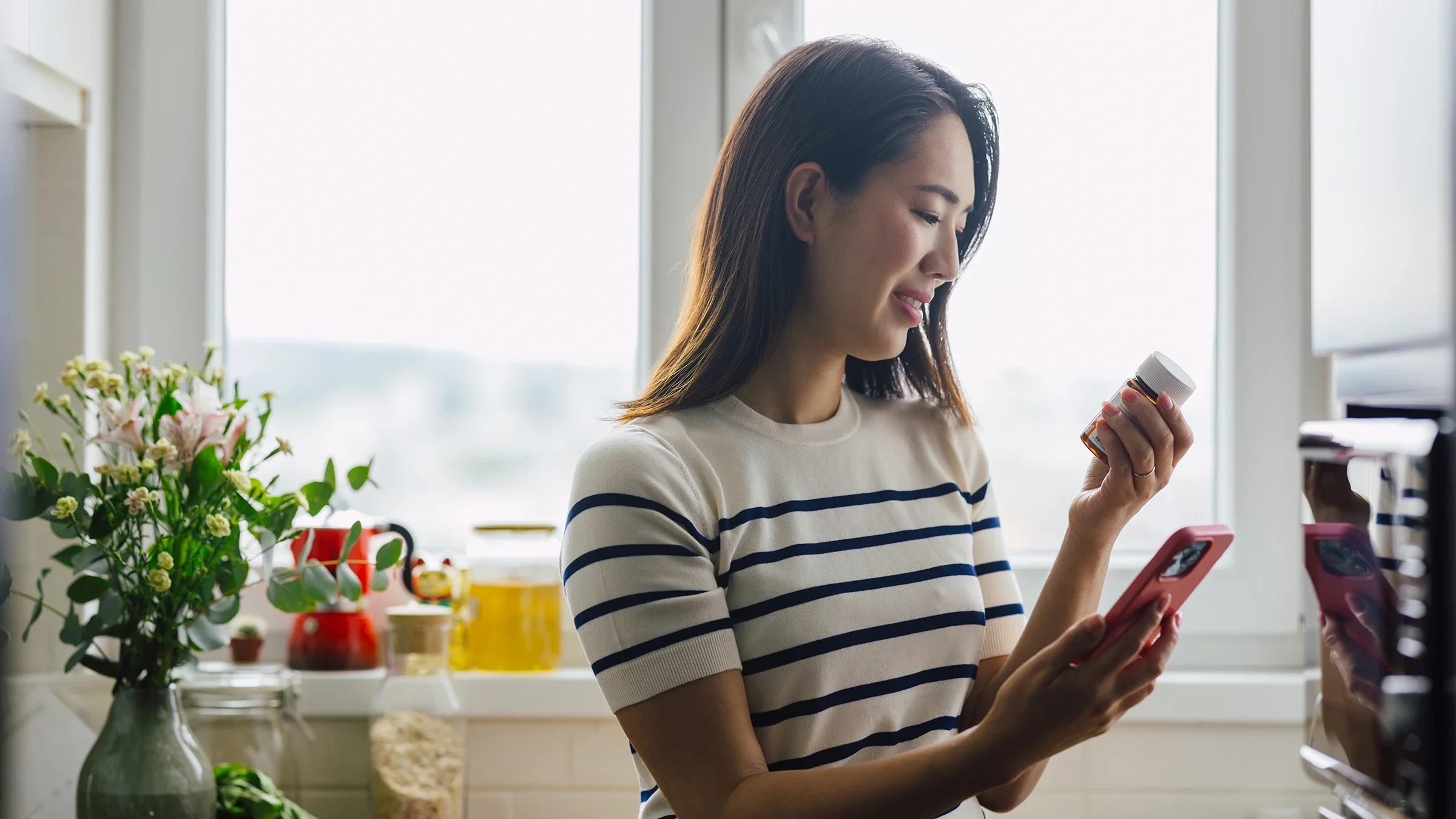 A woman is holding a pill bottle and looking up the medication on her smartphone.