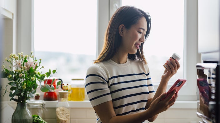A woman is holding a pill bottle and looking up the medication on her smartphone.