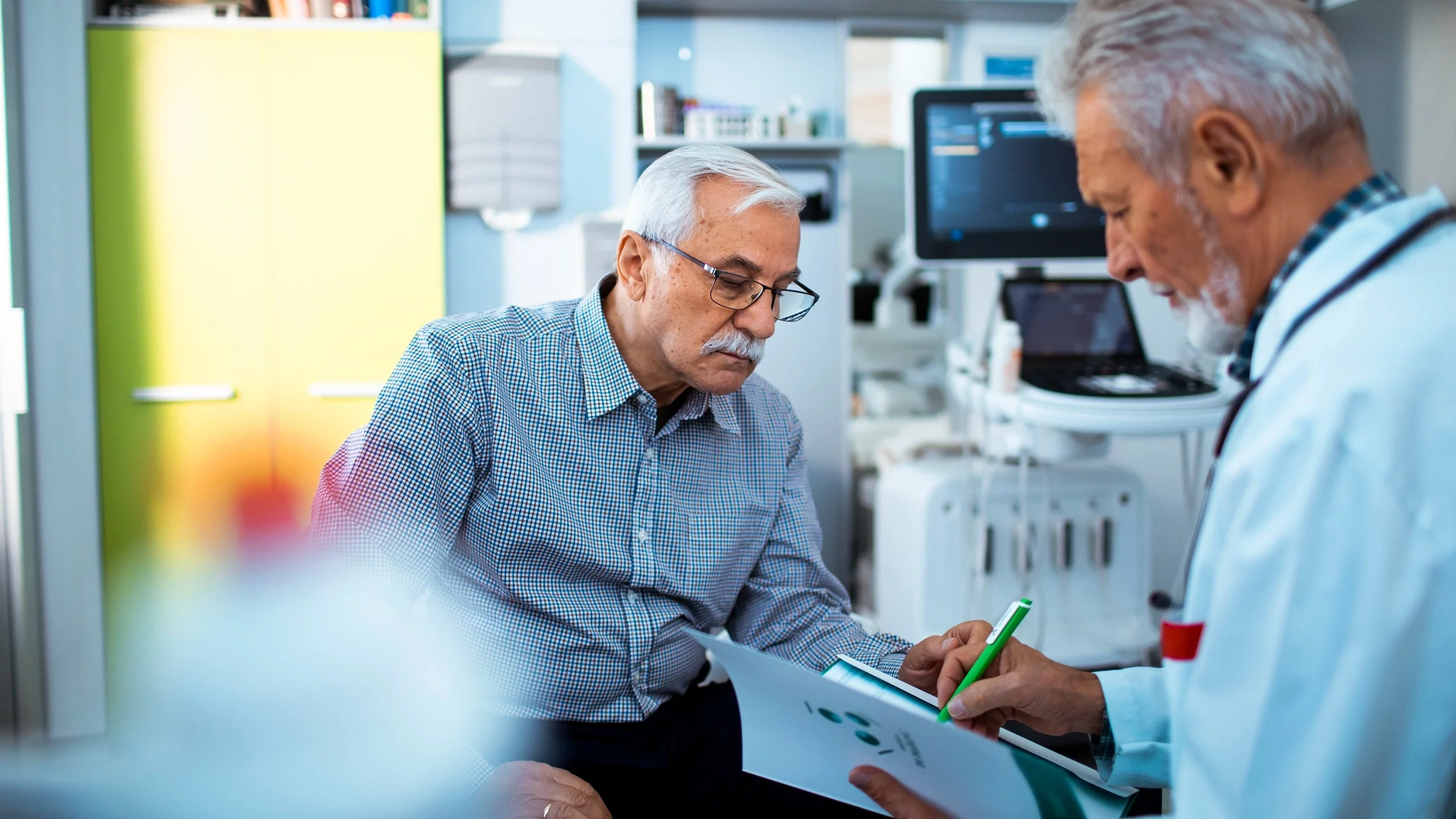 Older man at a consultation with his doctor. The doctor is taking notes while the patient looks at the paper the doctor is writing on.