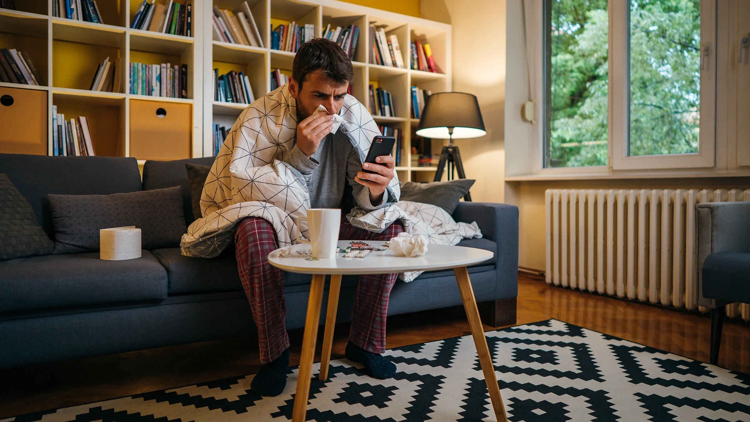Wrapped in a blanket, a man blows his nose as he sits on a sofa.