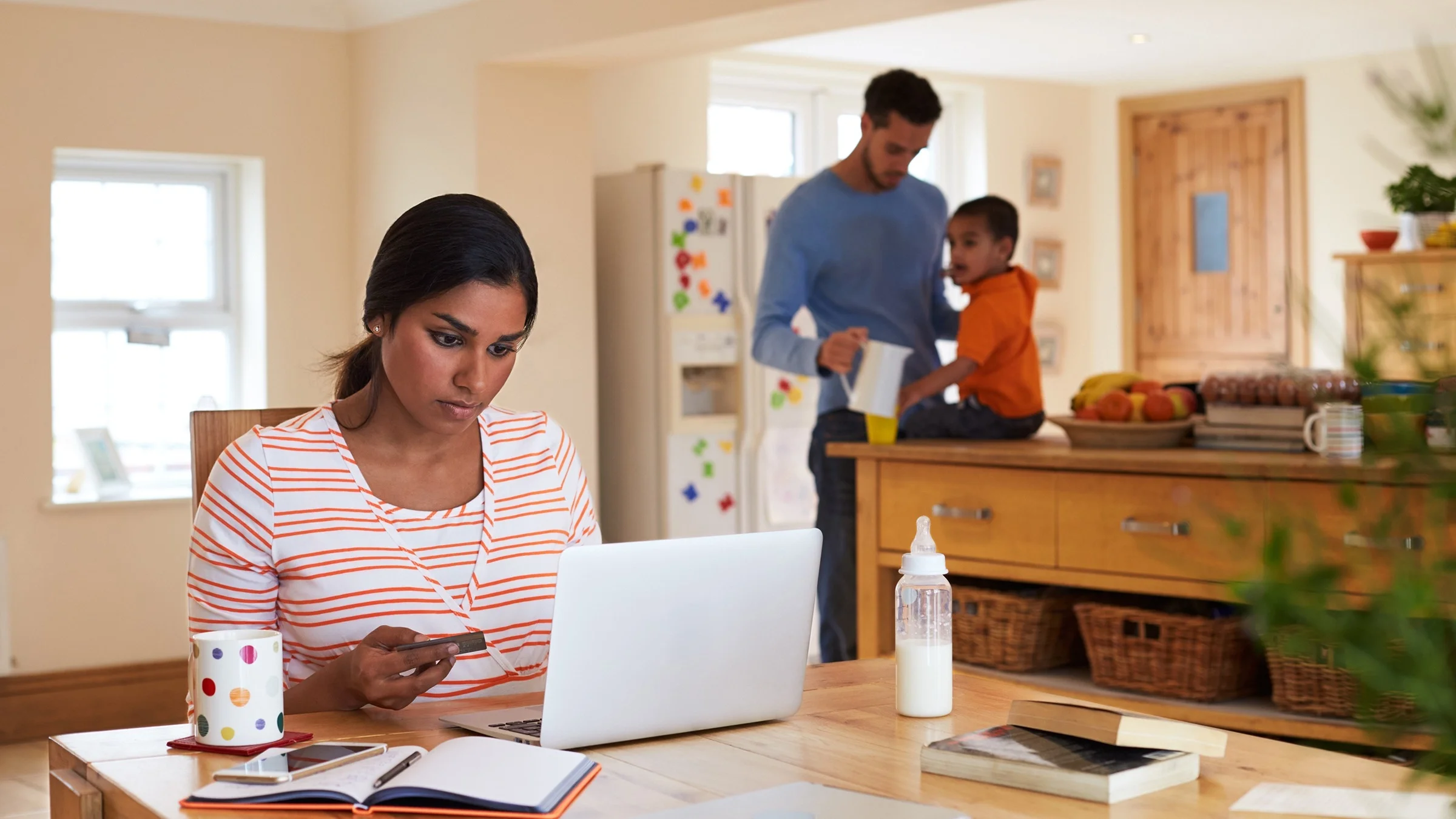 Young mother using her credit card to pay bills online. She is sitting at the kitchen table with her laptop while her partner and child are in the background in the kitchen.