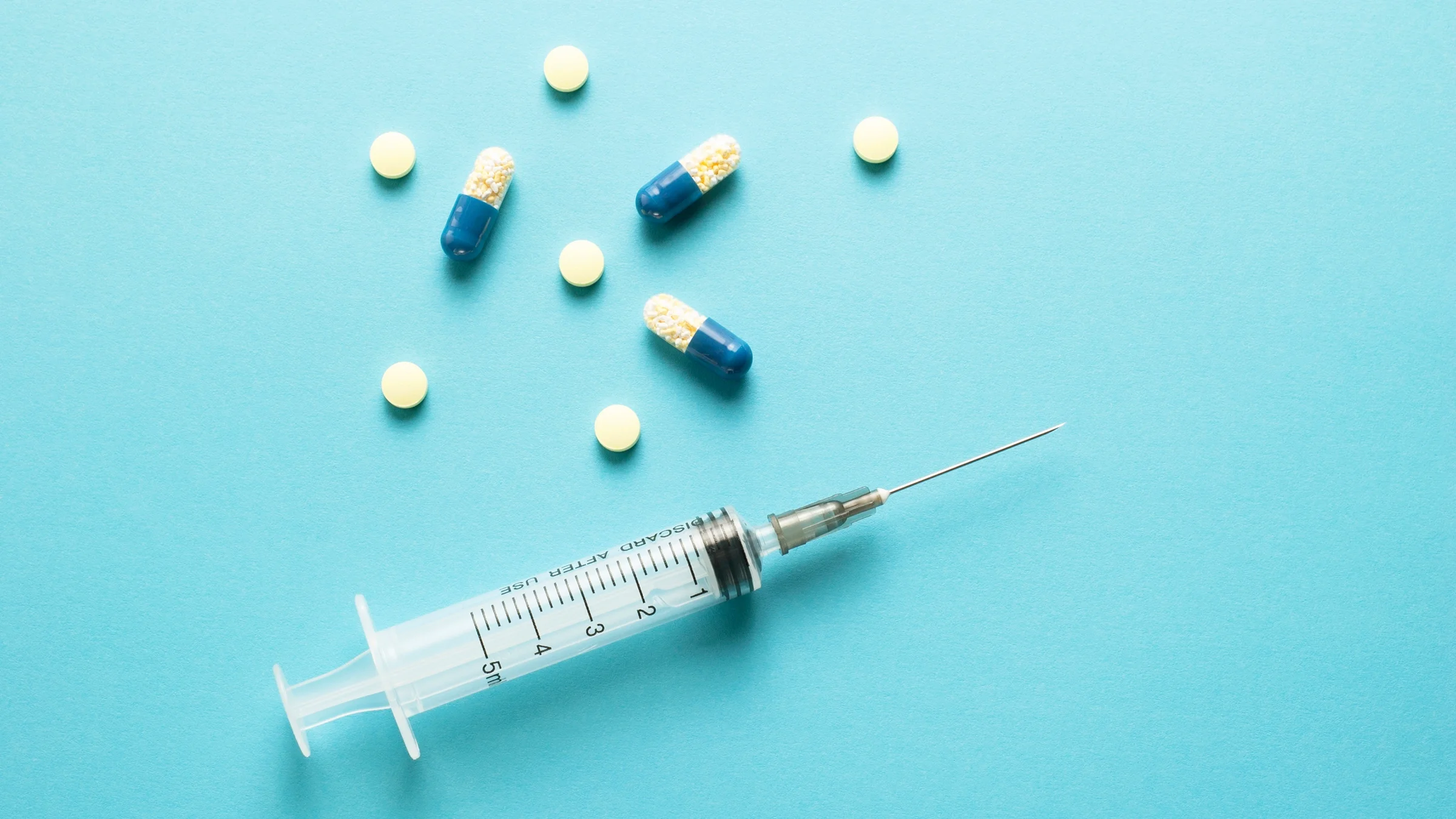 Syringe and pills on a teal background. There is a mix of tiny round white pills and blue and white oblong capsule pills.