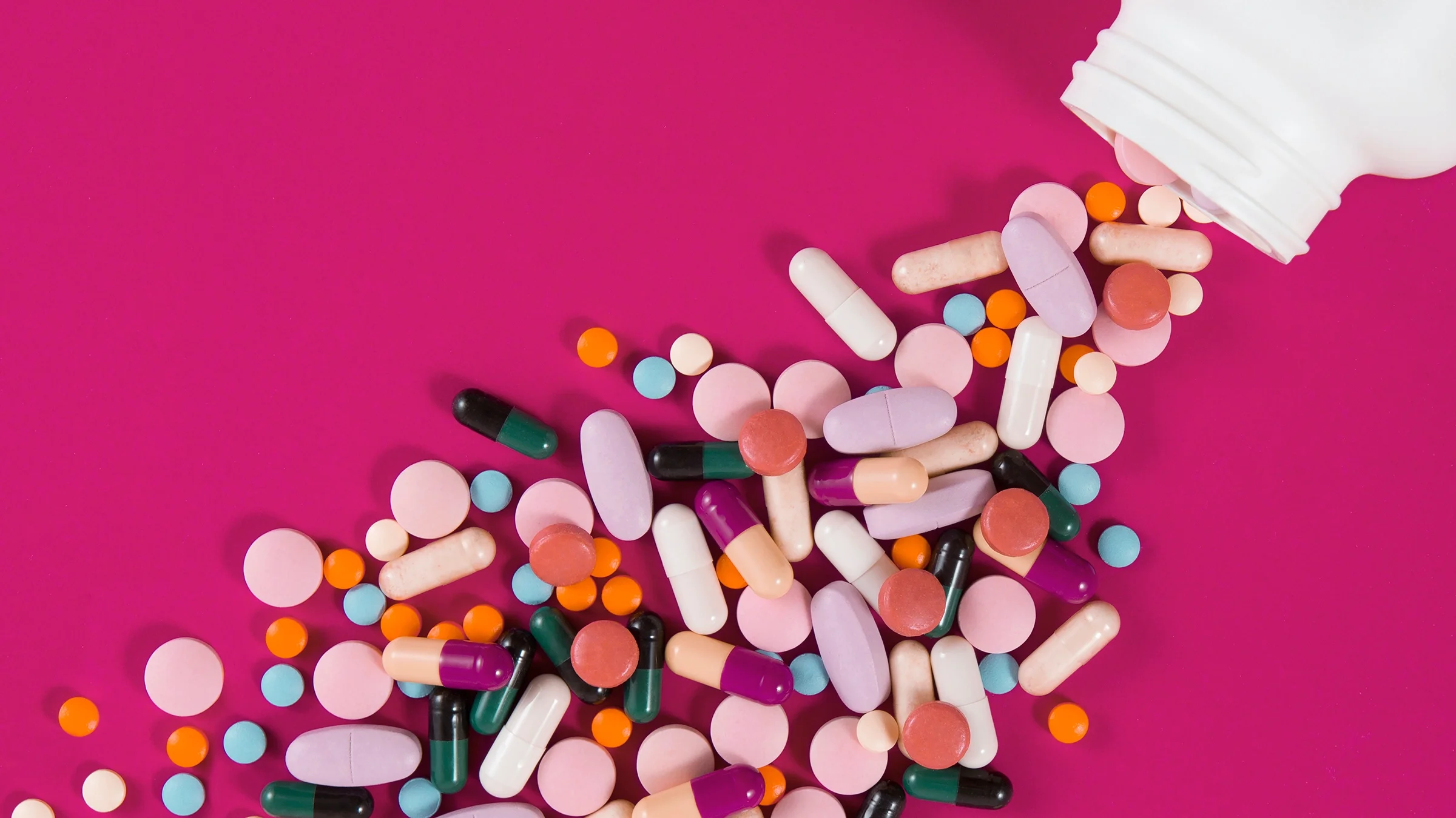 Various pills spilling out of white bottle on a pink background.