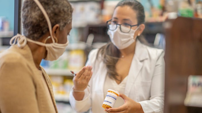 Older woman with short hair and white face mask on talking with the pharmacist through the plexiglass counter divider. They are reviewing one of her prescriptions.