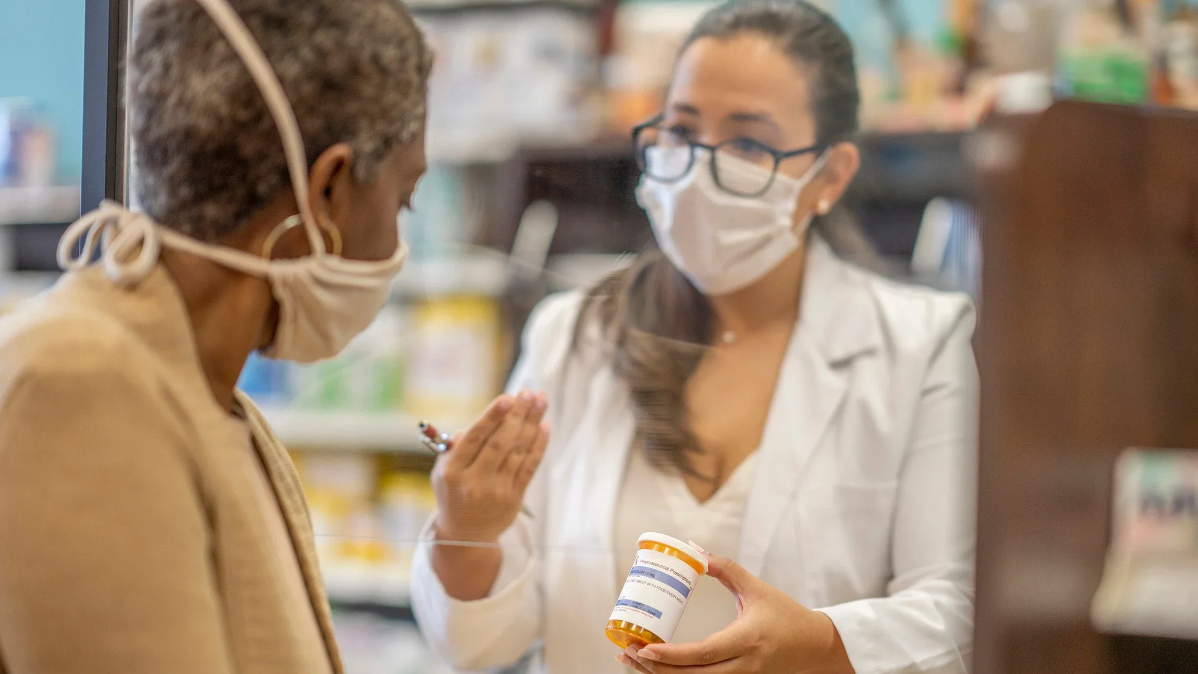 Older woman with short hair and white face mask on talking with the pharmacist through the plexiglass counter divider. They are reviewing one of her prescriptions.
FatCamera/E+ via Getty Images
