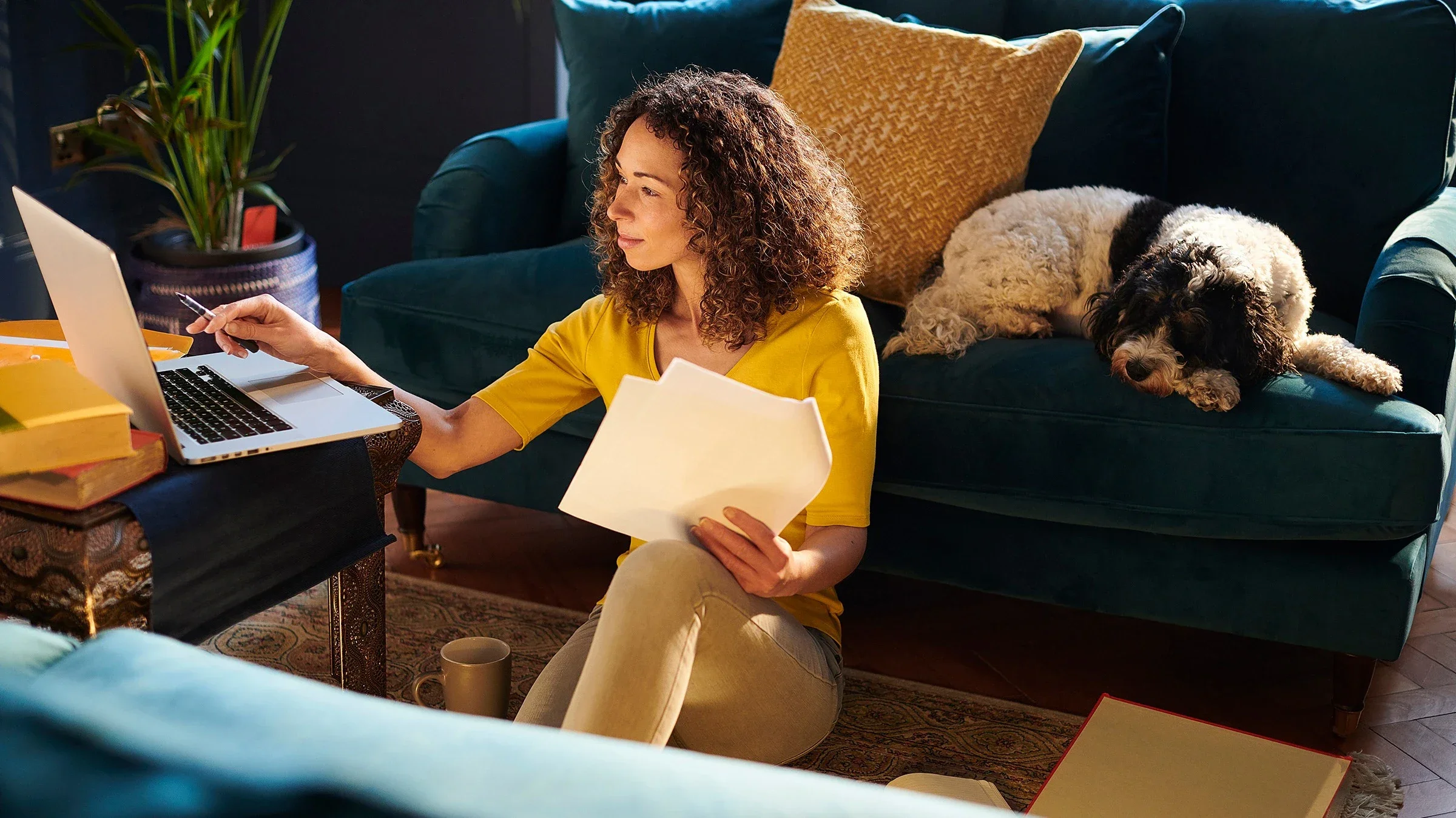 Woman sitting on her living room floor going over medical paper work and working on her laptop. He fluffy black and white dog is on the couch behind her.
sturti/E+ via Getty Images