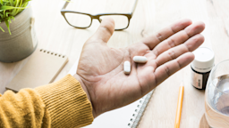 Open palm with two white pills with a desk background. There is a bottle of medicine and a water glass on the desk as well.
HAKINMHAN/iStock via Getty Images