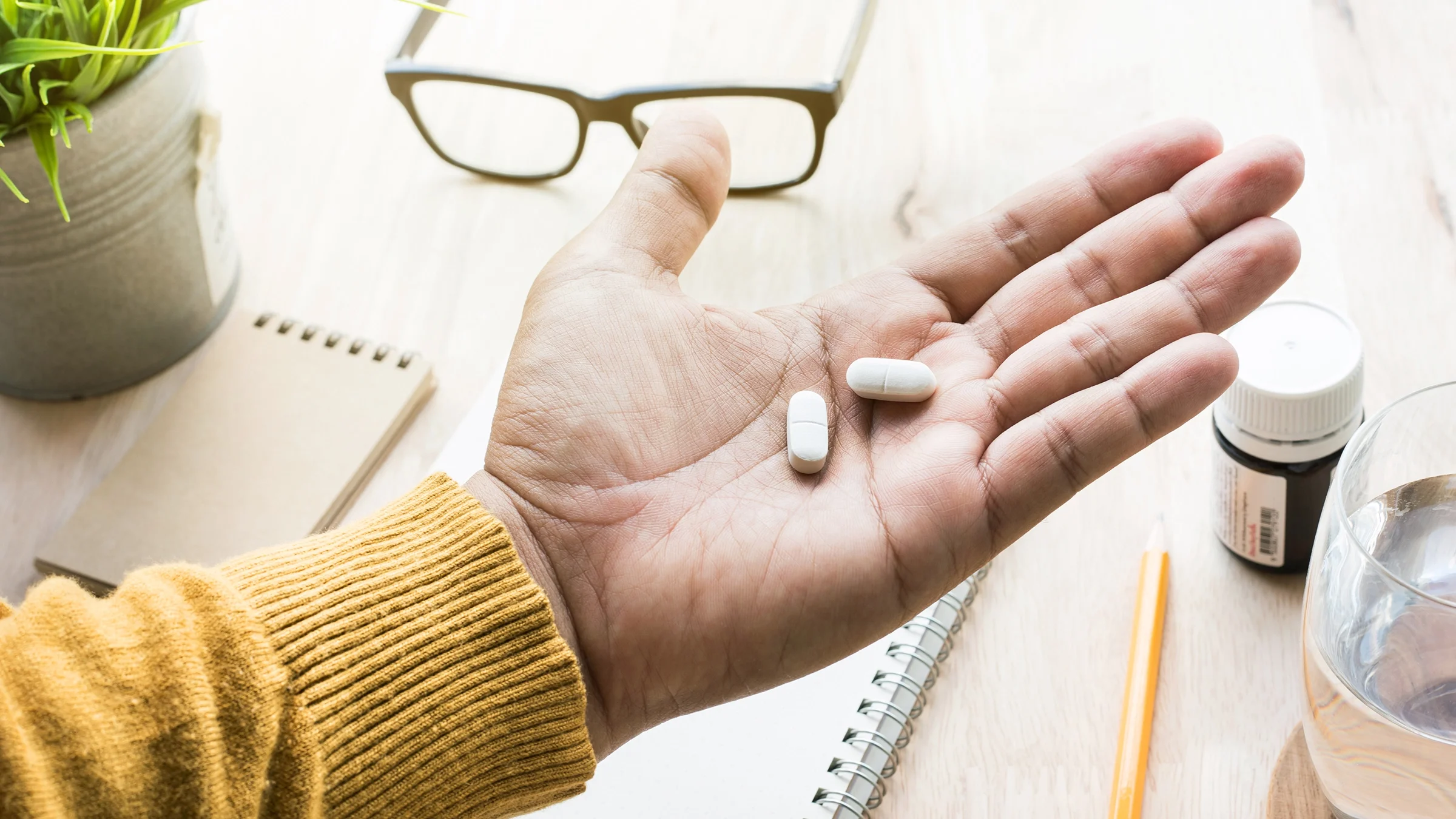 Open palm with two white pills with a desk background. There is a bottle of medicine and a water glass on the desk as well.