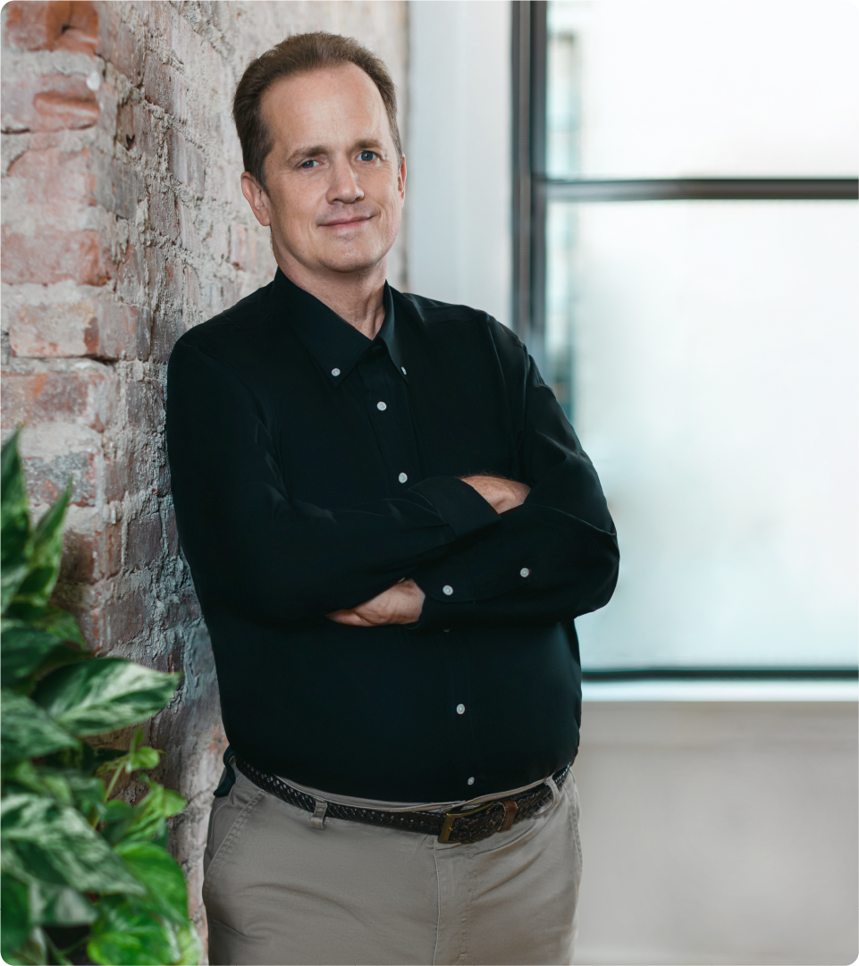 Portrait of Roger in a studio space with a brick wall and plant. 
