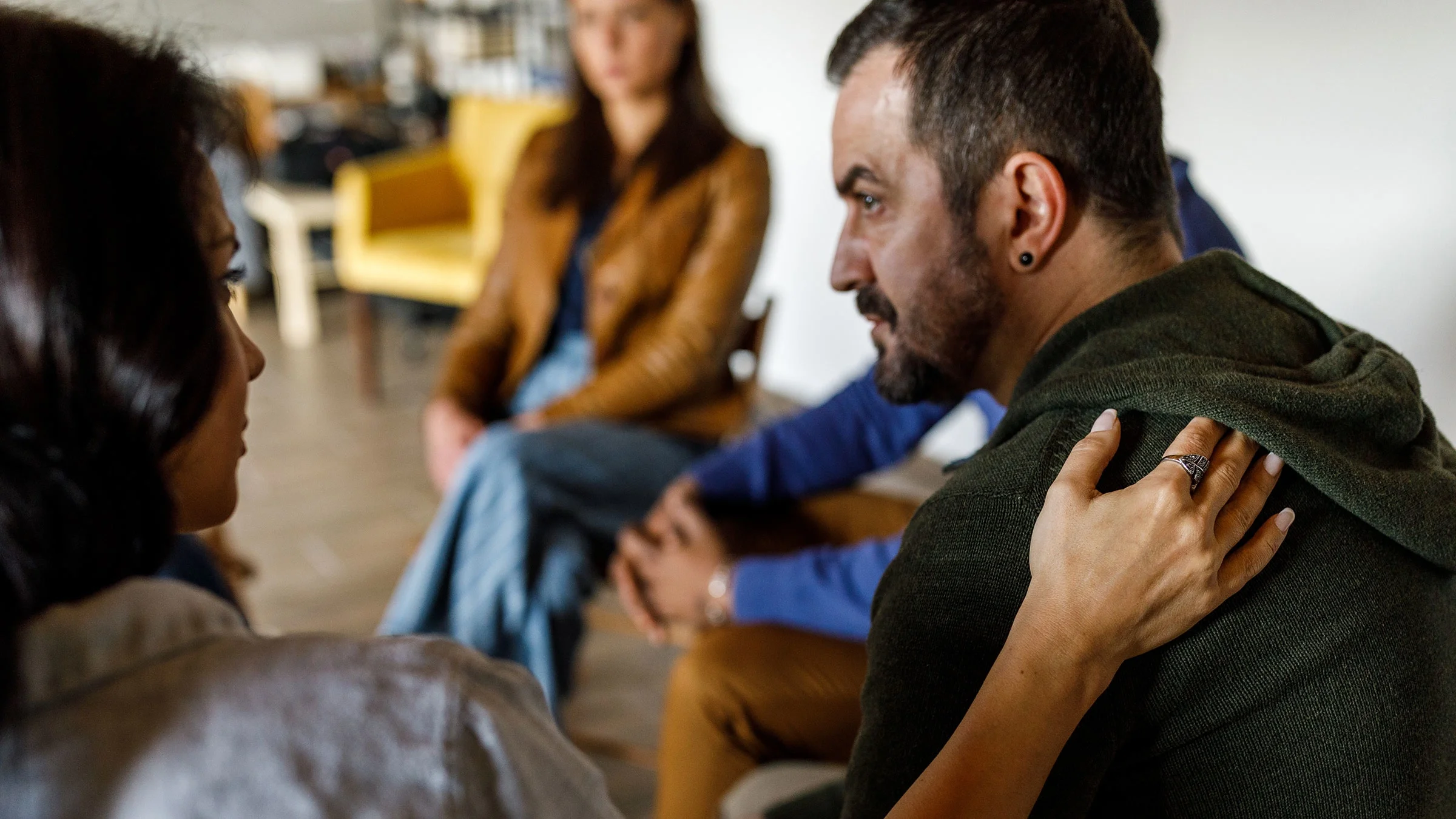 A compassionate woman puts a hand on a man's shoulder during a group therapy session.