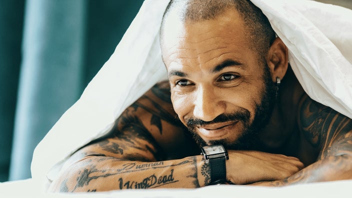 Close-up of a middle aged man with tattoos under the white sheets of his bed. He is resting his head on his hands.