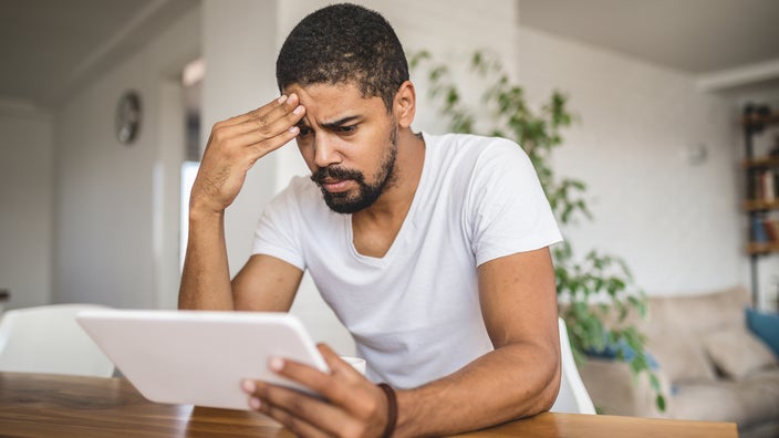 Young man reading the news on his tablet at home. He has his hand on his forehead in distress.