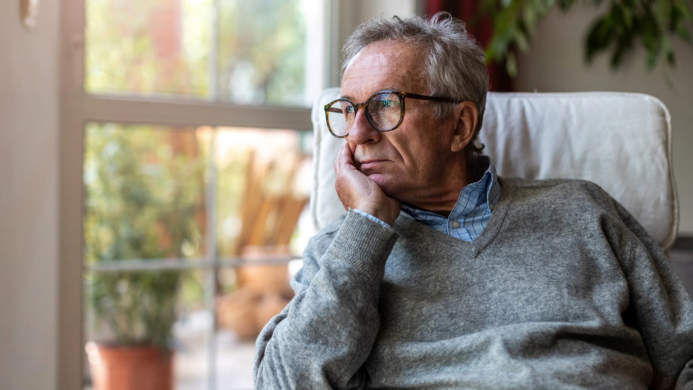 Older man in glasses and a sweater sitting in a living room chair looking out the window.