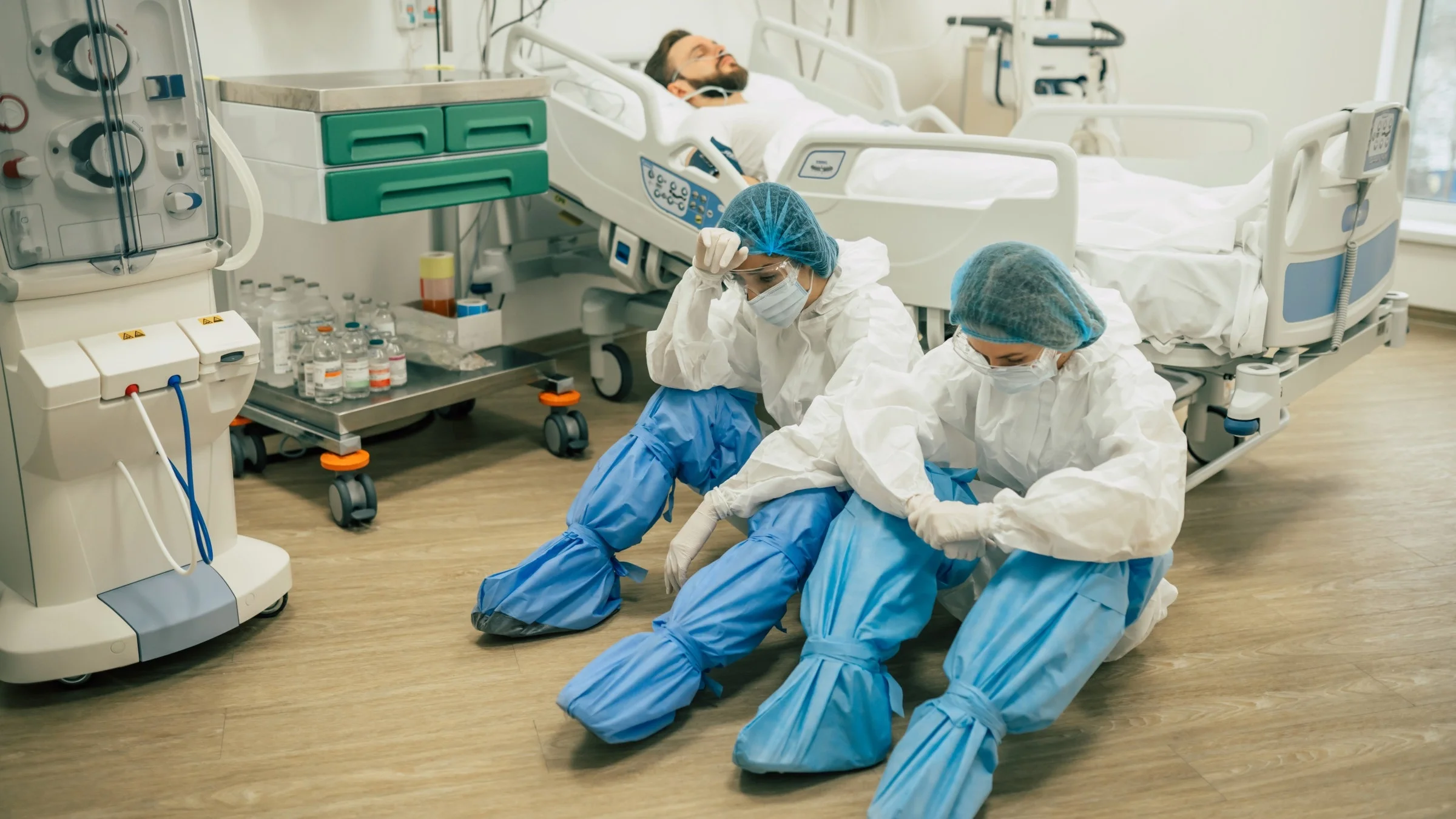 Two exhaused doctors sit on the floor next to a patients hospital bed in full PPE while the patient is on oxygen.
