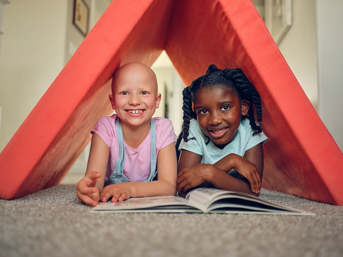 Little girl with alopecia playing and reading a book with her friend in a fort they created from a red padded mat.