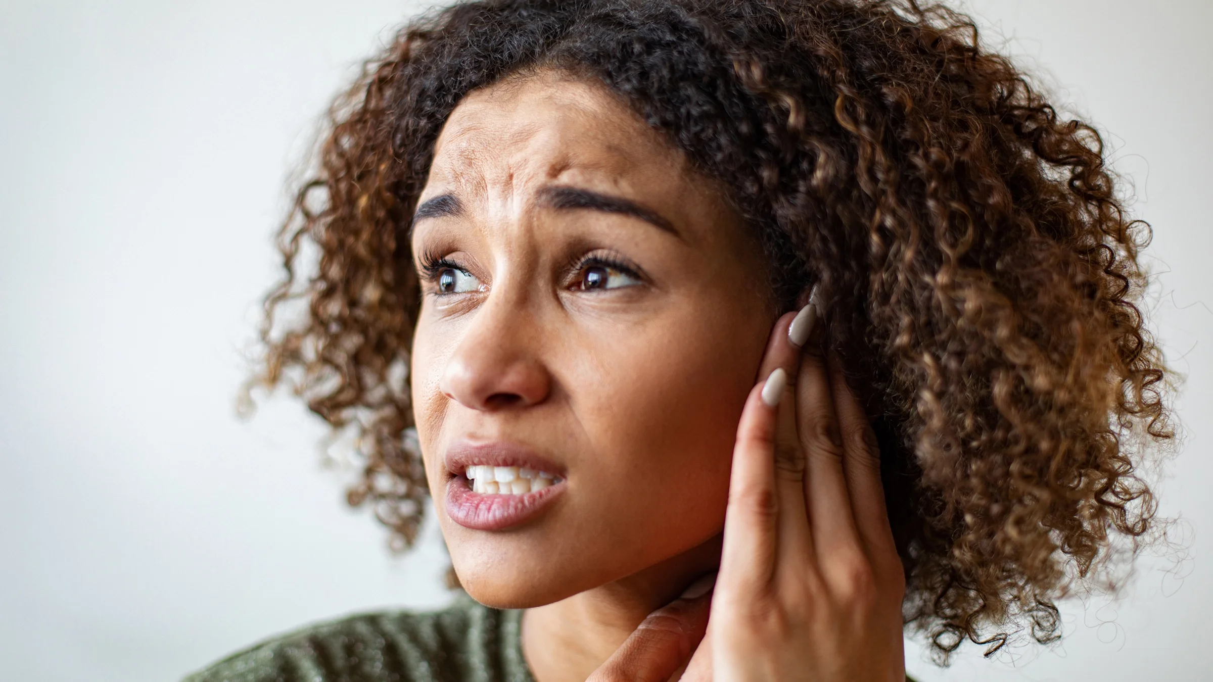 Close-up portrait of a woman with curly hair. She is wincing and rubbing her ear in pain.
