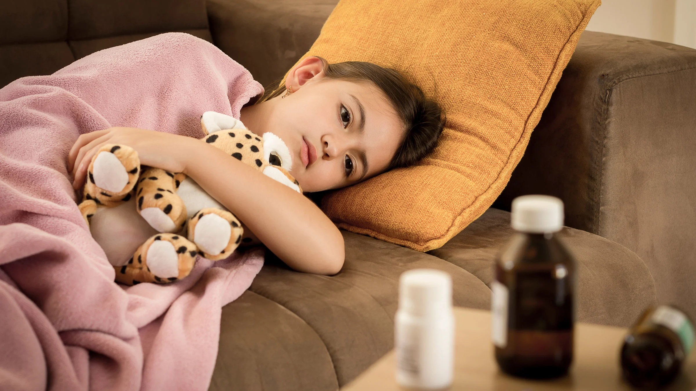 A sick child curled up in a blanket on a couch, hugging a stuffed animal. There is medicine on the table in front of them.