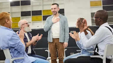 Man standing up to speak to AA group circle. They are applauding him.
SeventyFour/iStock via Getty Images