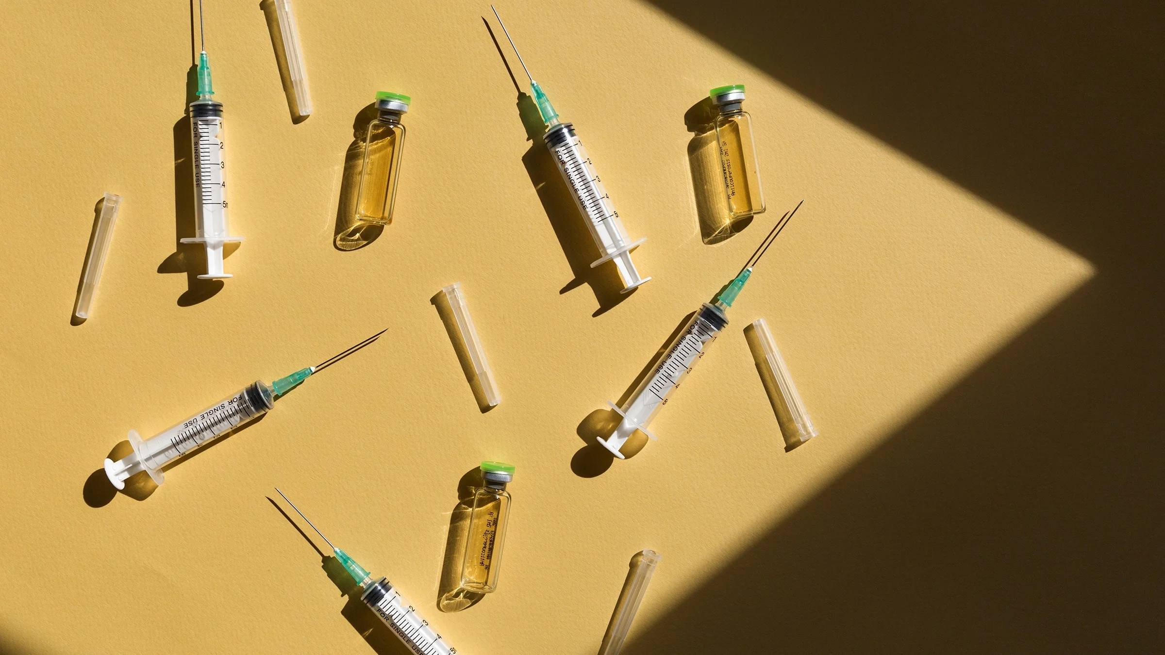 Flaylay of syringes and vaccine vials on a yellow shadow-y background.