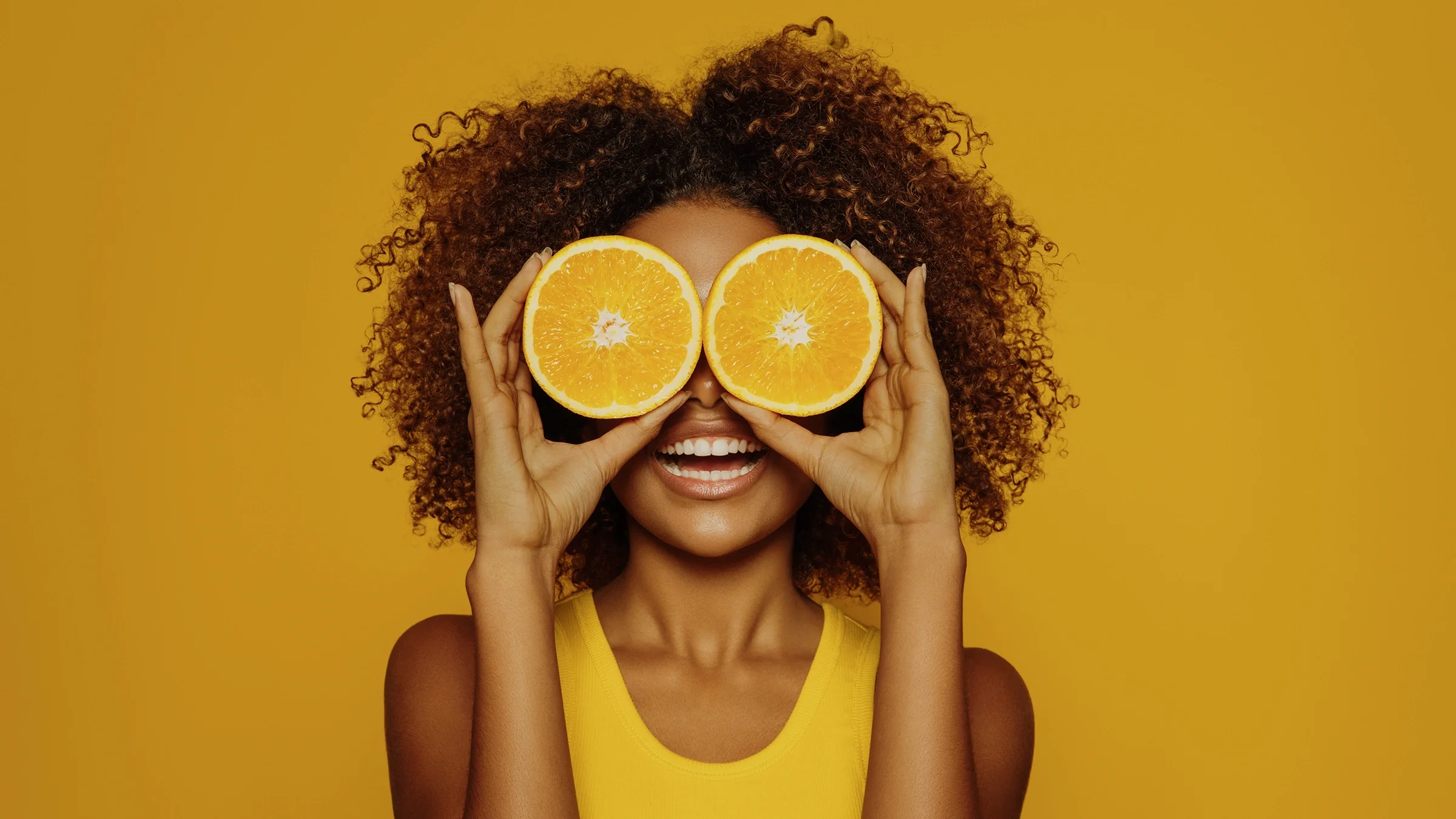 Woman with curly hair holding orange halves up to her eyes on an orange studio background.