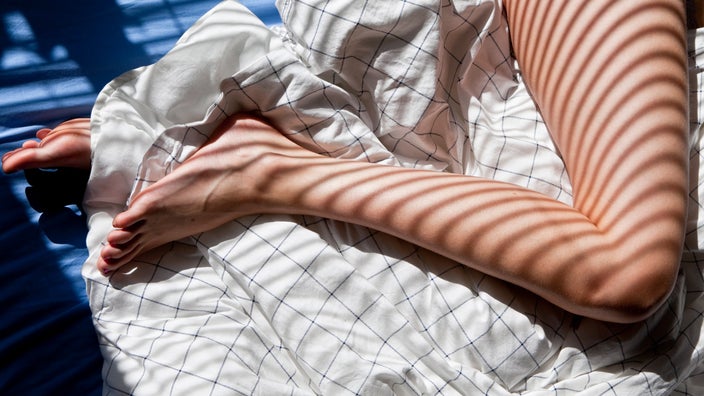 Cropped shot of a woman's legs intertwined in bed sheets from the hip down with the shadow of the blinds on her.