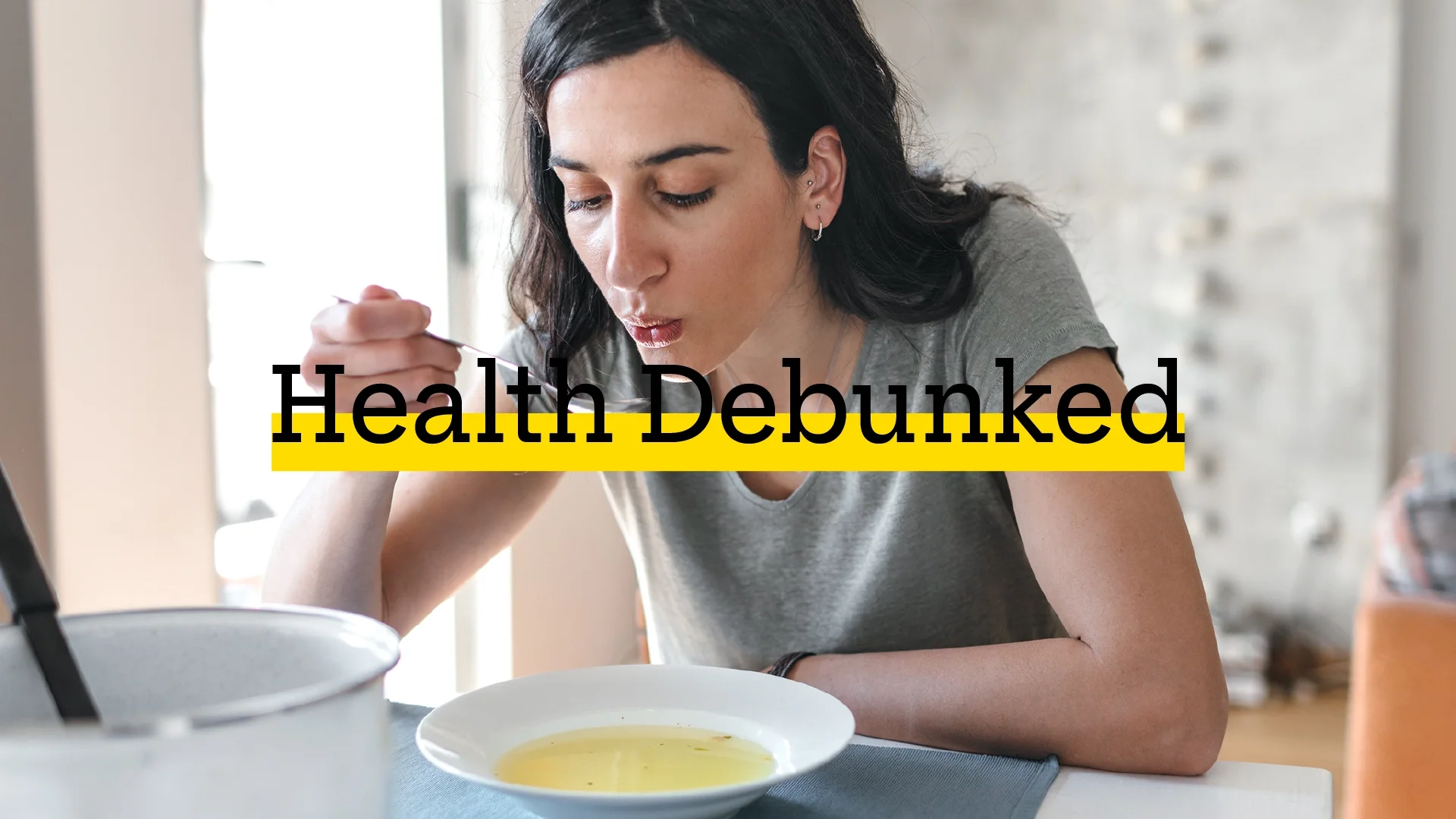 Woman with black hair eating chicken noodle soup. She is blowing on the spoonful of soup to cool it down. There is added text on top reading “Health Debunked” with a yellow highlight bar behind it.