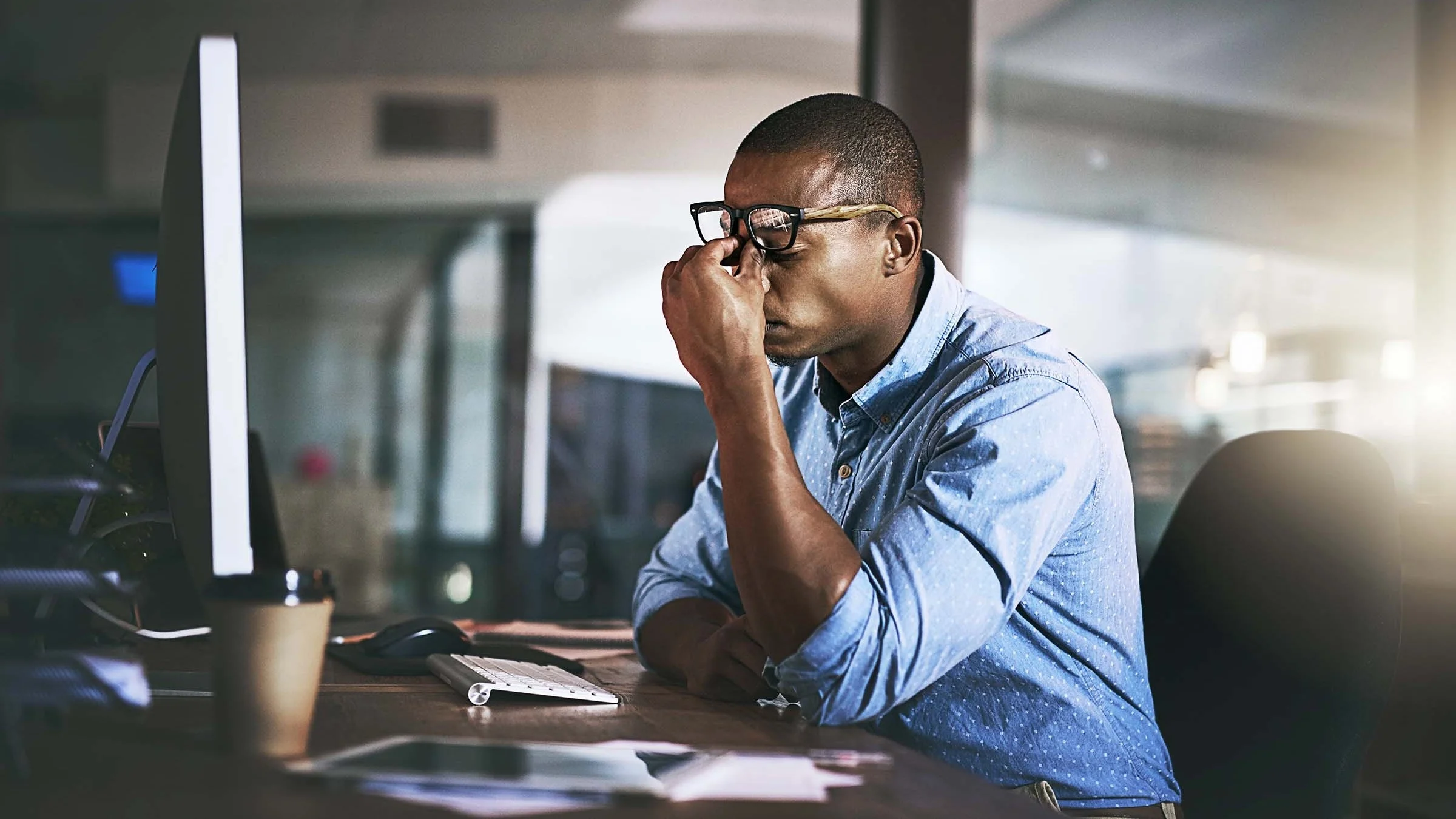 Young man sitting at his desk at work. He is pinching his nose with pointer finger and thumb while raising his glasses up.