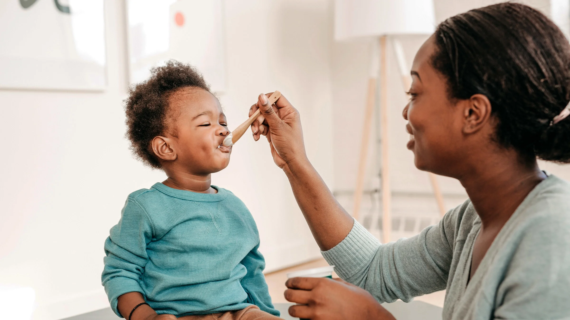 Mother feeding her toddler yogurt with a wooden spoon.