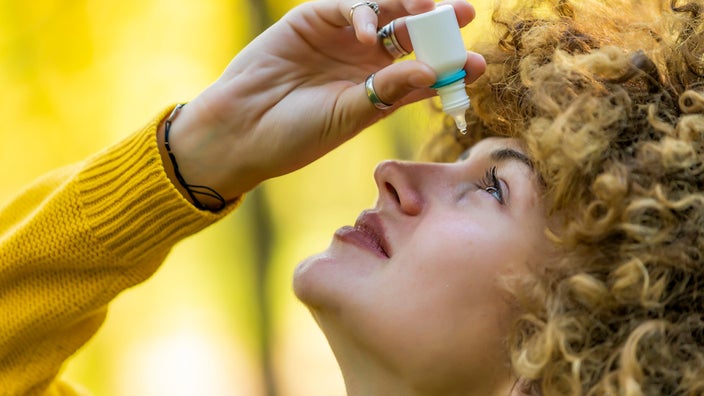 Close-up of a woman with curly hair putting eye drops in her eye. She is outside with a blurry yellow green background.
