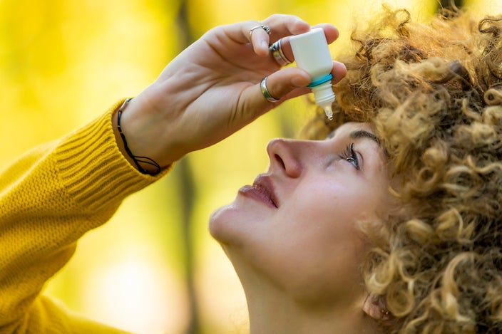 Close-up of a woman with curly hair putting eye drops in her eye. She is outside with a blurry yellow green background.