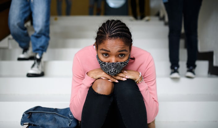 Portrait of a teenage girl sitting on the steps of her high school. She is wearing a mask and resting her chin on her knees. She looks slightly sad.