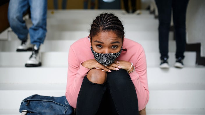 Portrait of a teenage girl sitting on the steps of her high school. She is wearing a mask and resting her chin on her knees. She looks slightly sad.