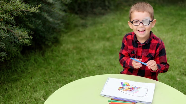 Little boy with glasses on and one eye covered, sitting at a play table outside and drawing with coloring pens.
