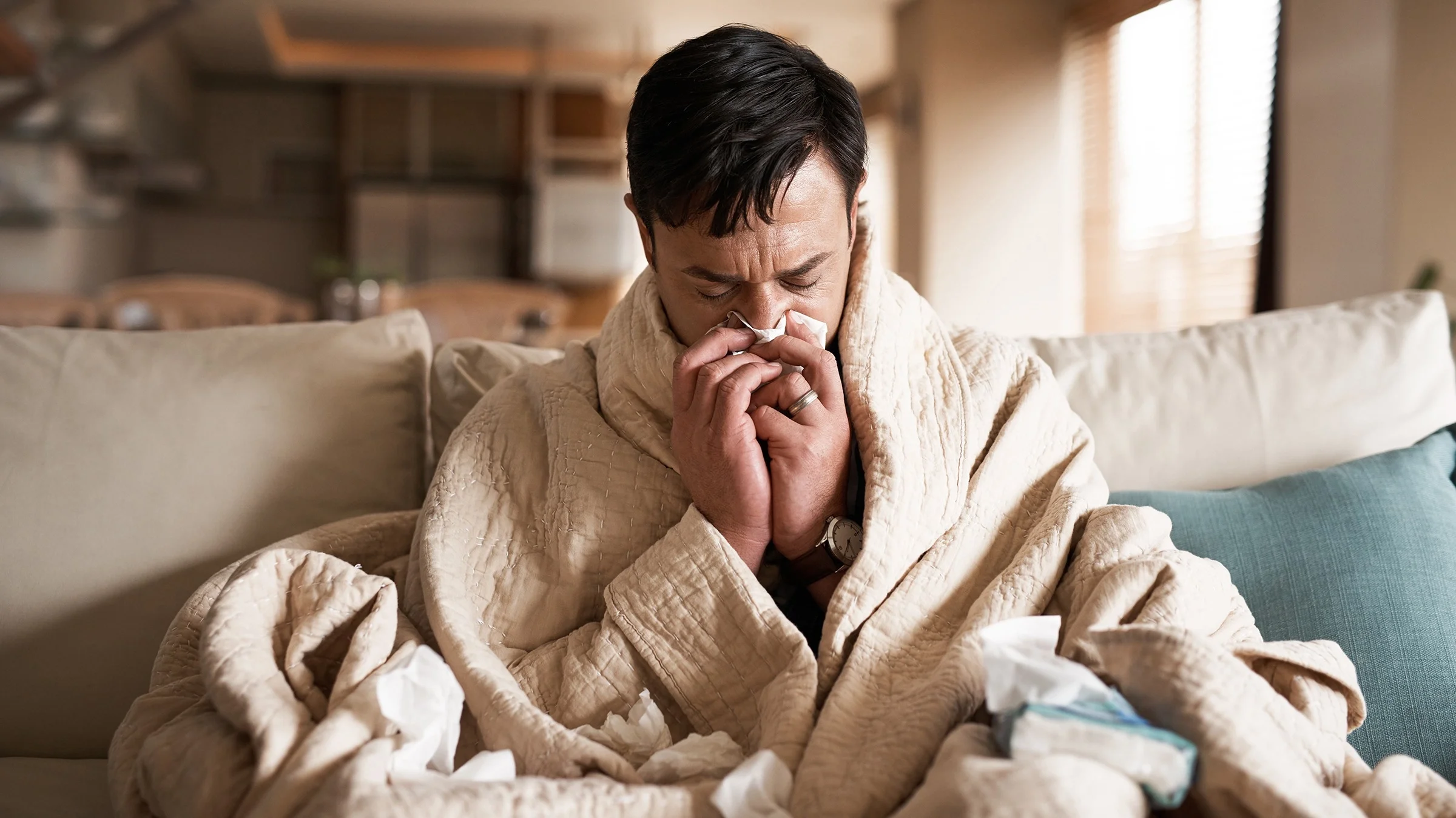 Man sitting on the couch wrapped up in a blanket blowing his nose with tissues surrounding him.