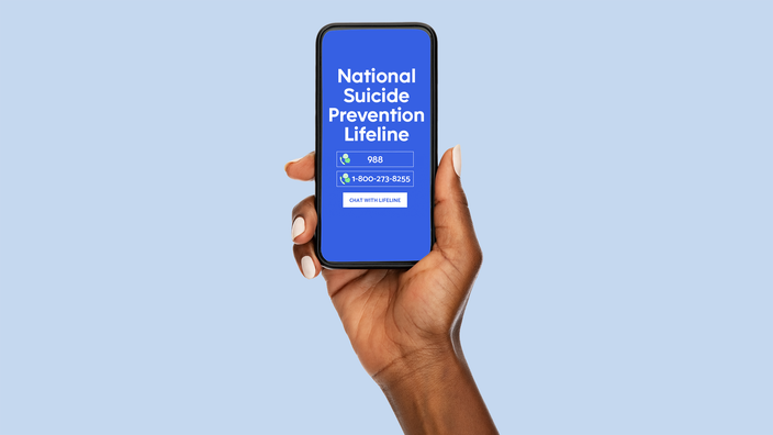Isolated hand holding smartphone on a light blue background. The phone screen displays a mock-up of the Suicide Prevention Hotline numbers to call.