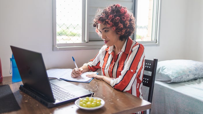 A woman is sitting at a desk and taking notes while looking at her computer.